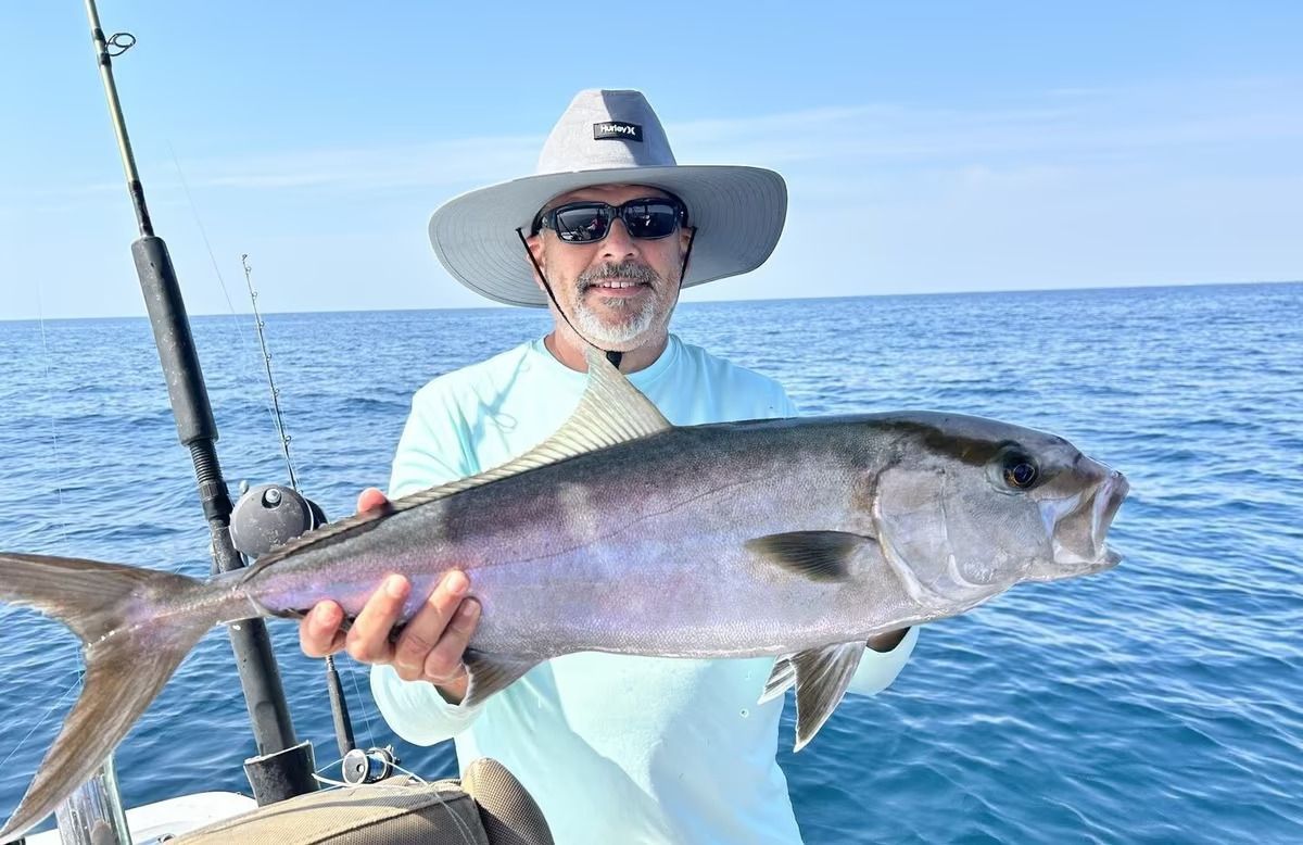 Man holding a large silver fish on a boat at sea, wearing a hat and sunglasses