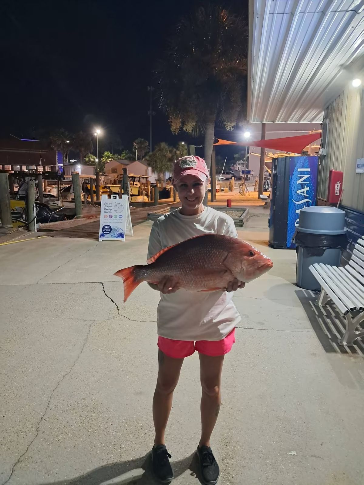 Person holding a large fish on a lit nighttime sidewalk near shops and outdoor seating