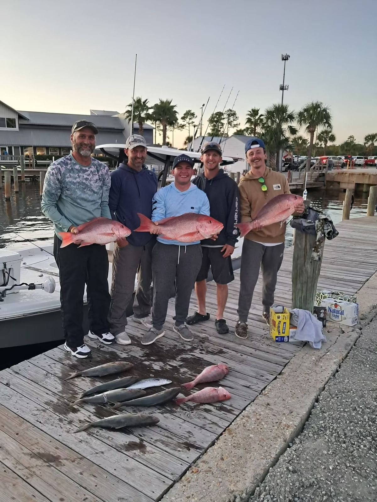 Five anglers hold large fish on a dock at sunset, with more fish laid out at their feet.