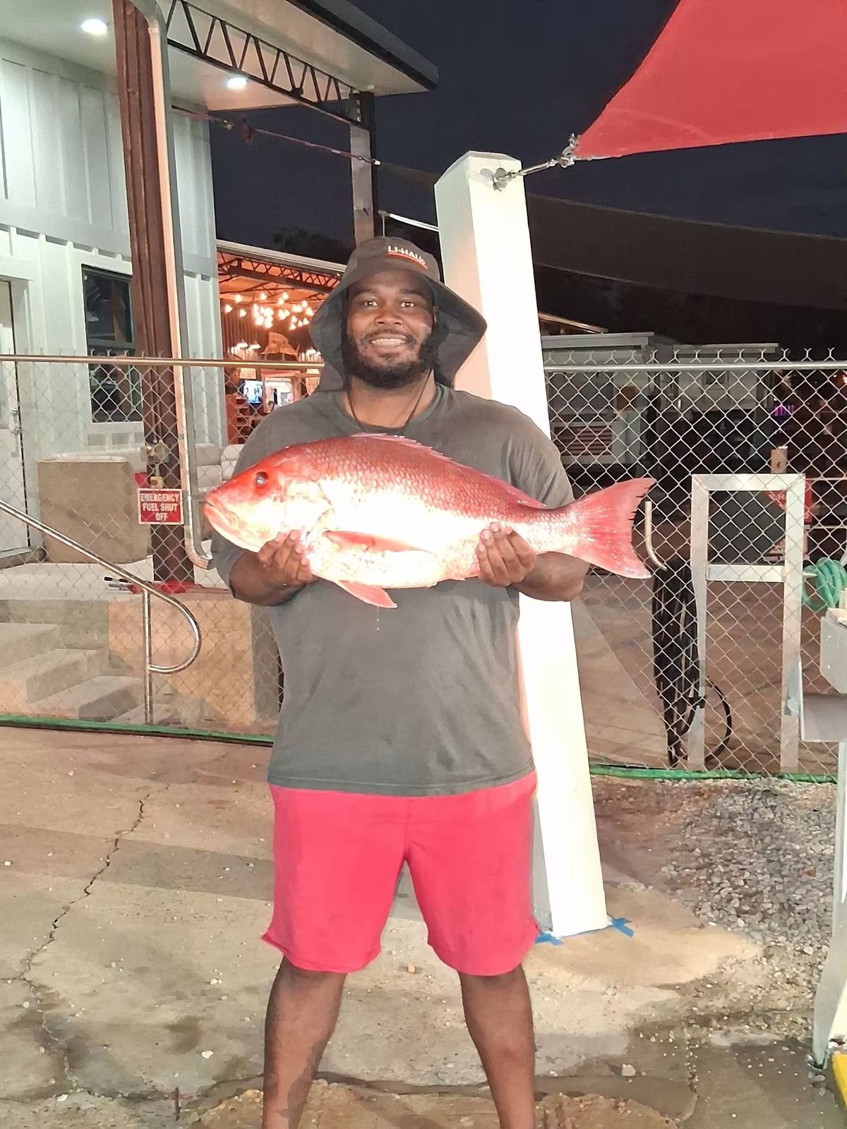 Man holding a large red fish outdoors at night, smiling in a red shirt and shorts