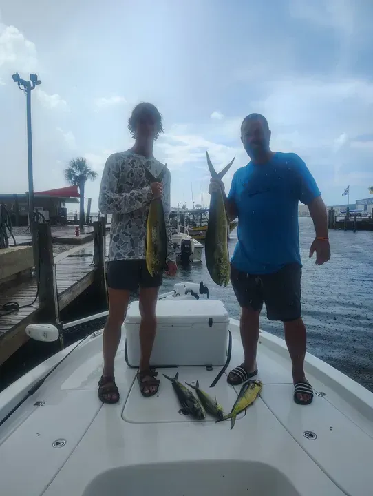 Two people standing on a boat dock, holding fish, with rods and gear on the deck.