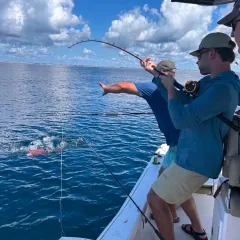 Two people fishing from a boat, one reeling in a bent rod over blue ocean under cloudy sky