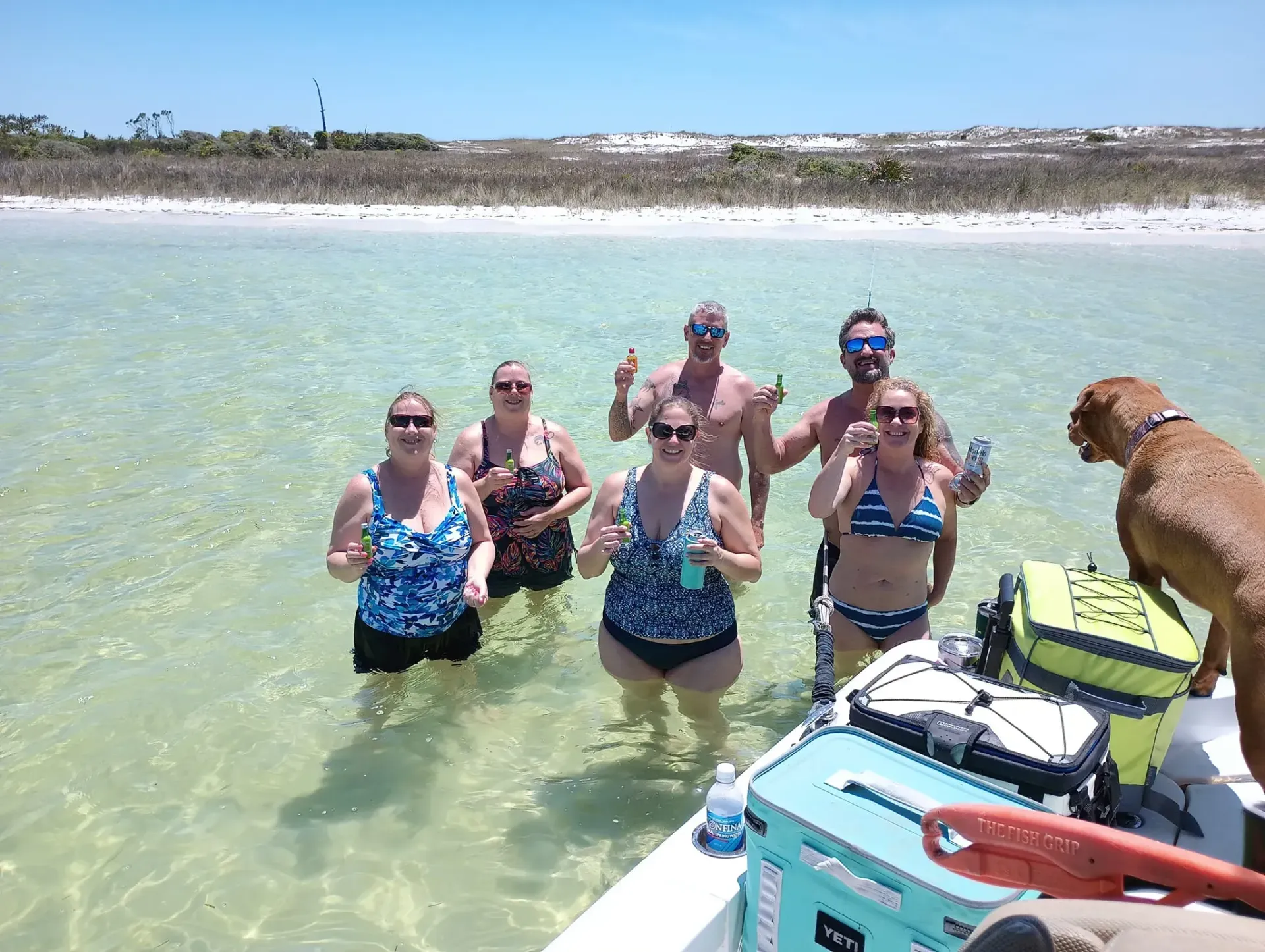 Group of people posing in shallow clear water at a beach, with a dog on a kayak in the foreground.
