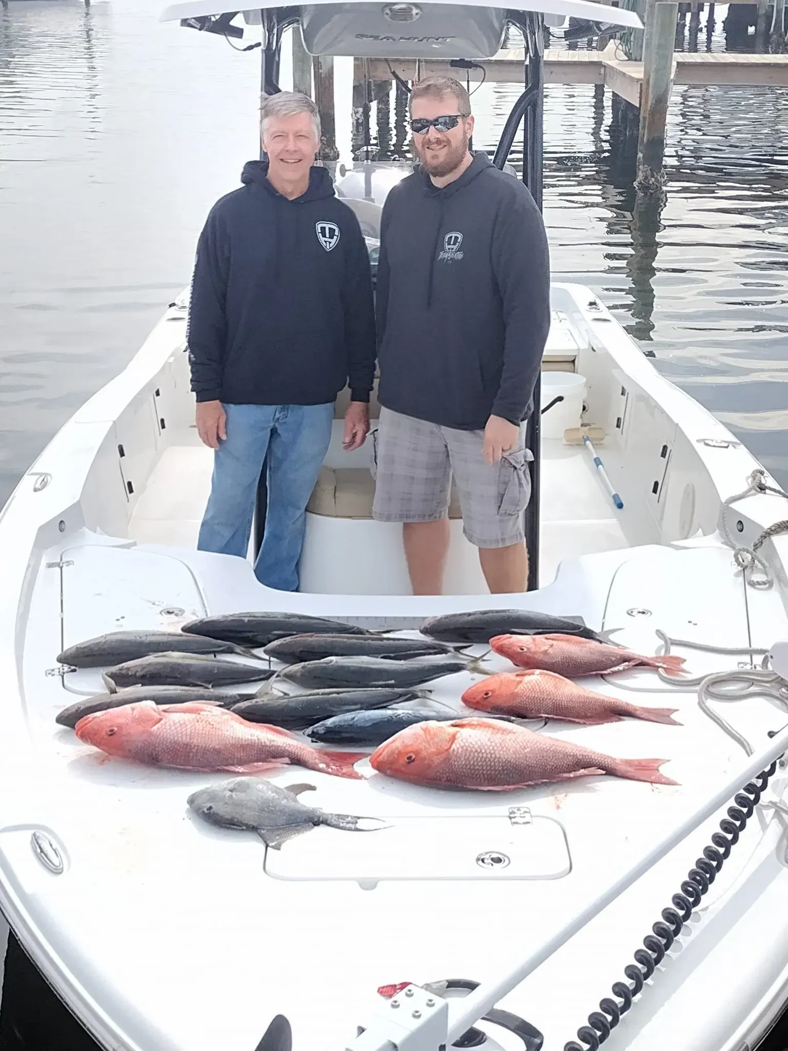 Two people on a boat with a large catch of fish laid out on the deck, dock in the background.