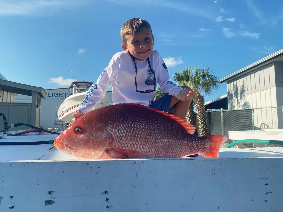 Person smiling behind a large red fish on a boat dock under a blue sky