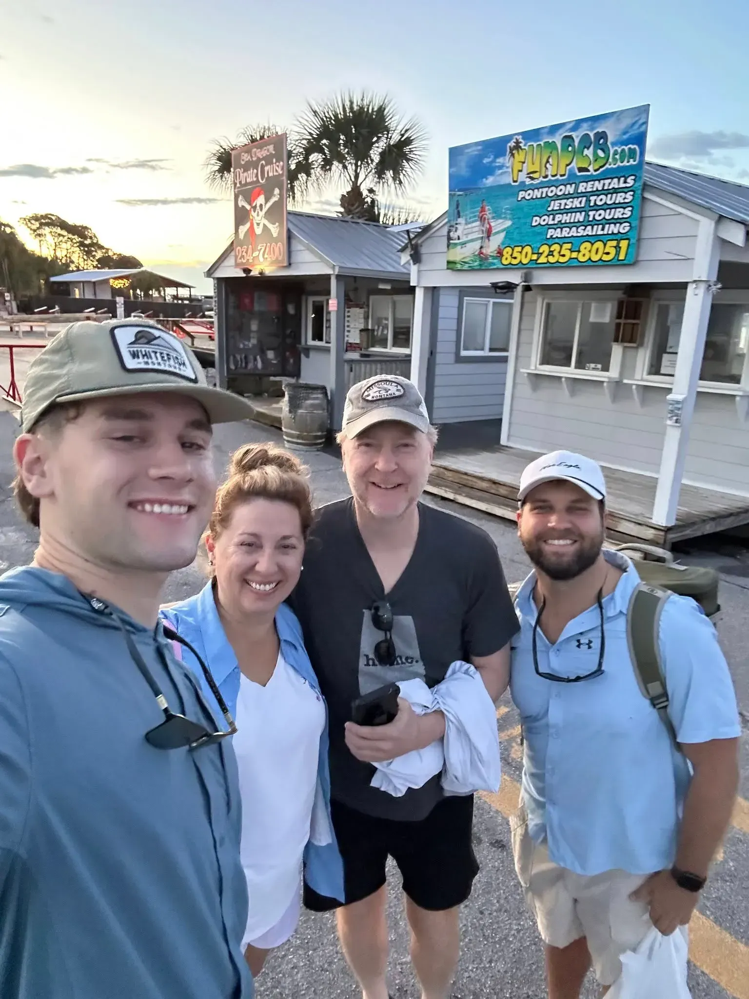 Four friends smiling in front of a beachside shop at sunset, holding a selfie camera.