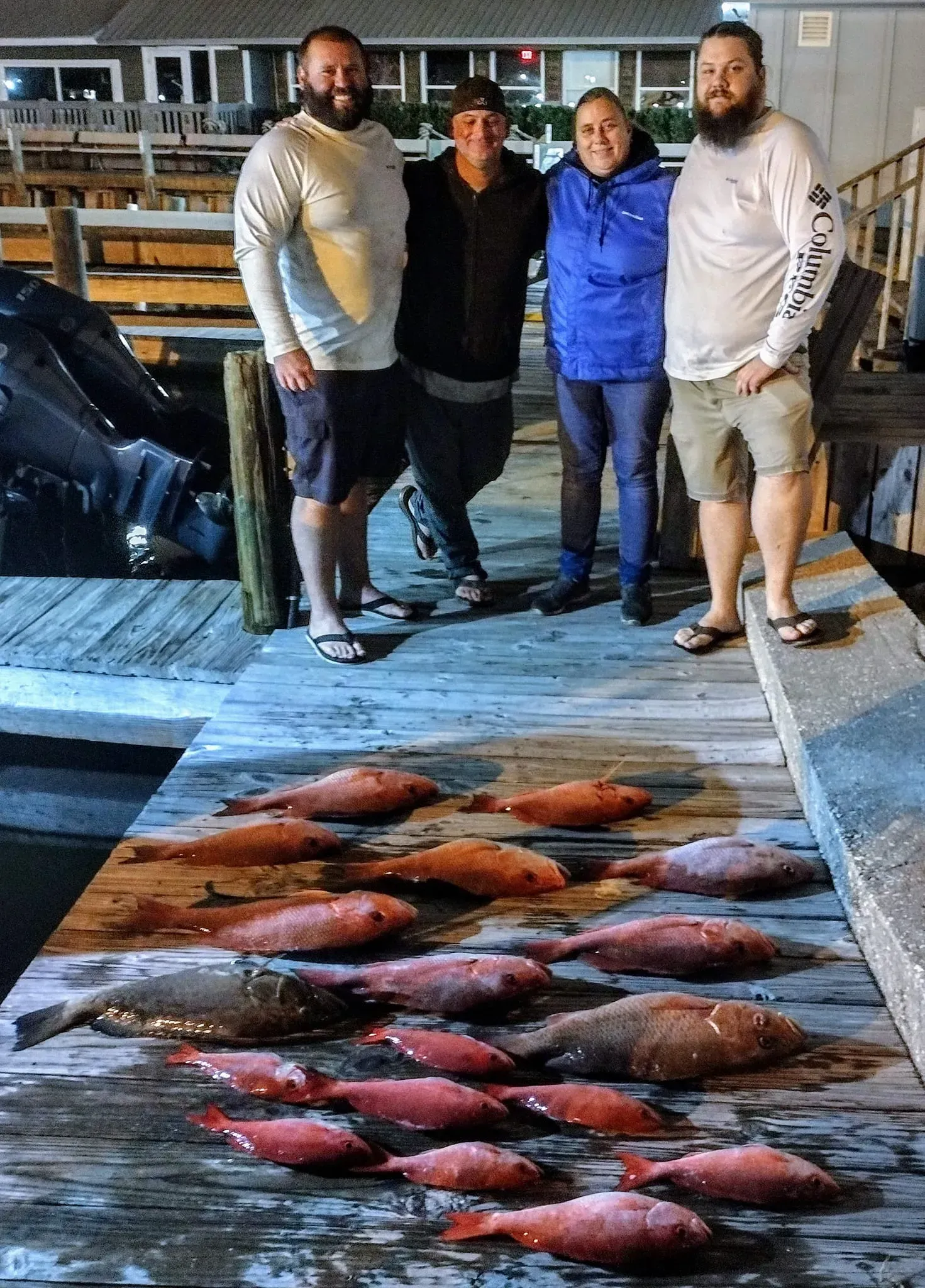 Four people stand on a dock beside a large pile of freshly caught fish at night.