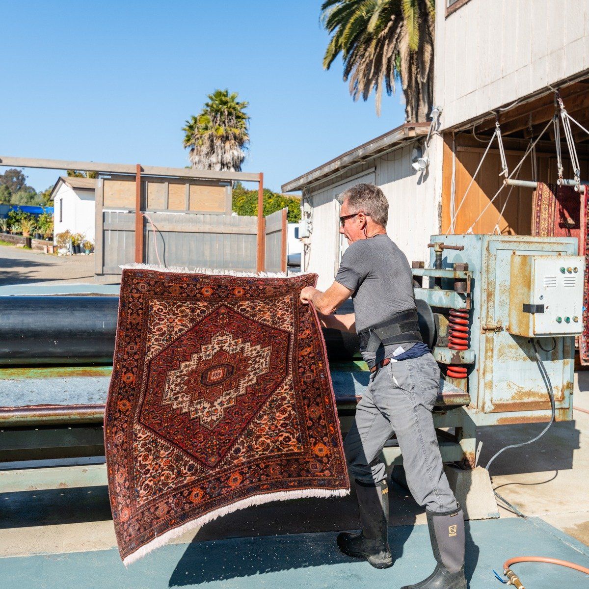 A person wearing a grey shirt and boots feeds a patterned, dark red rug into a large cleaning machine outdoors.