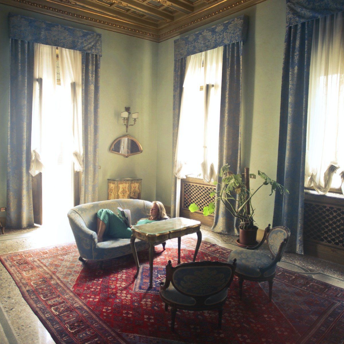 A person lounges on a light blue sofa in a sunlit, high-ceilinged room with ornate gold trim, curtains, and a red rug.