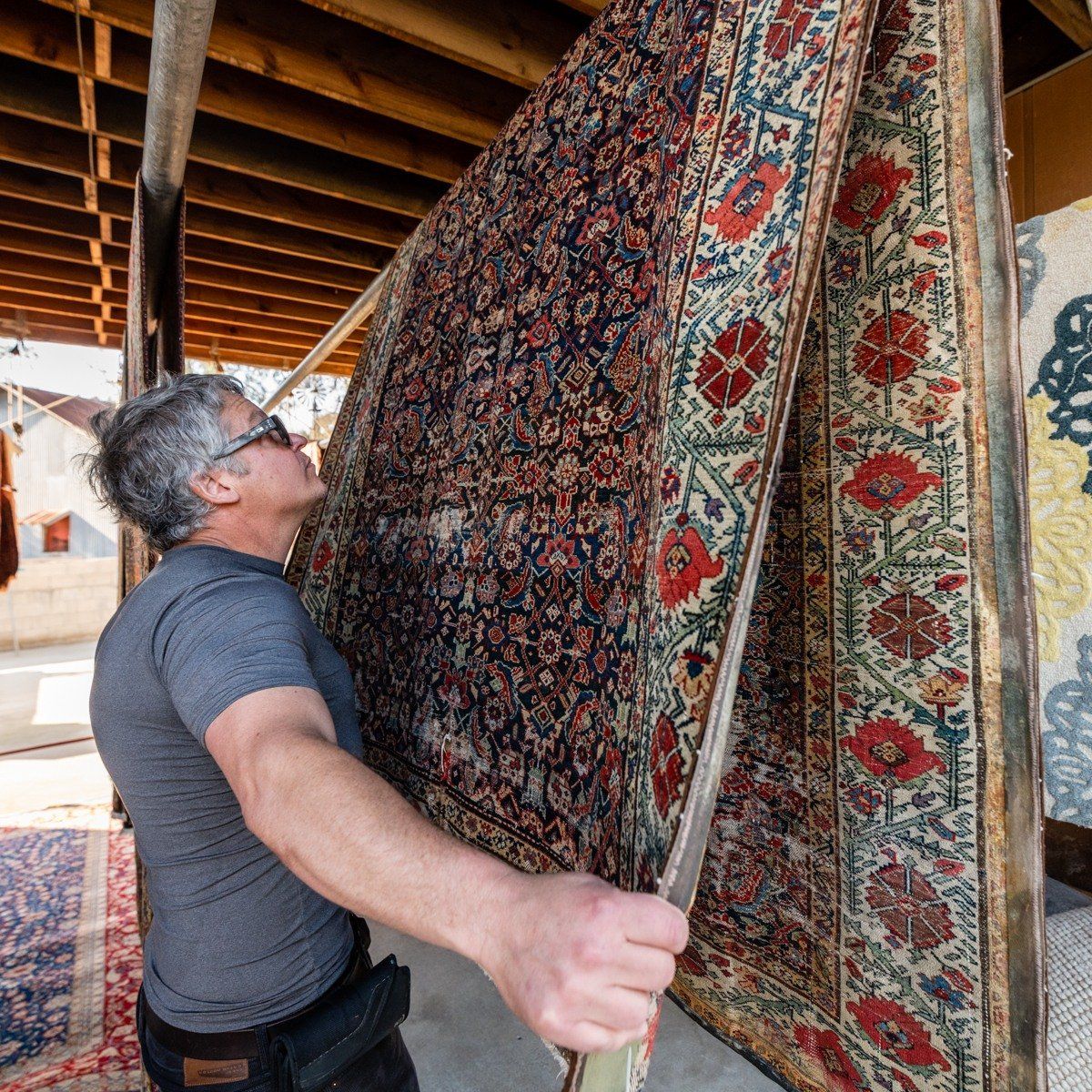A person examining a large, intricately patterned, dark-toned Persian-style rug hanging in an outdoor space.