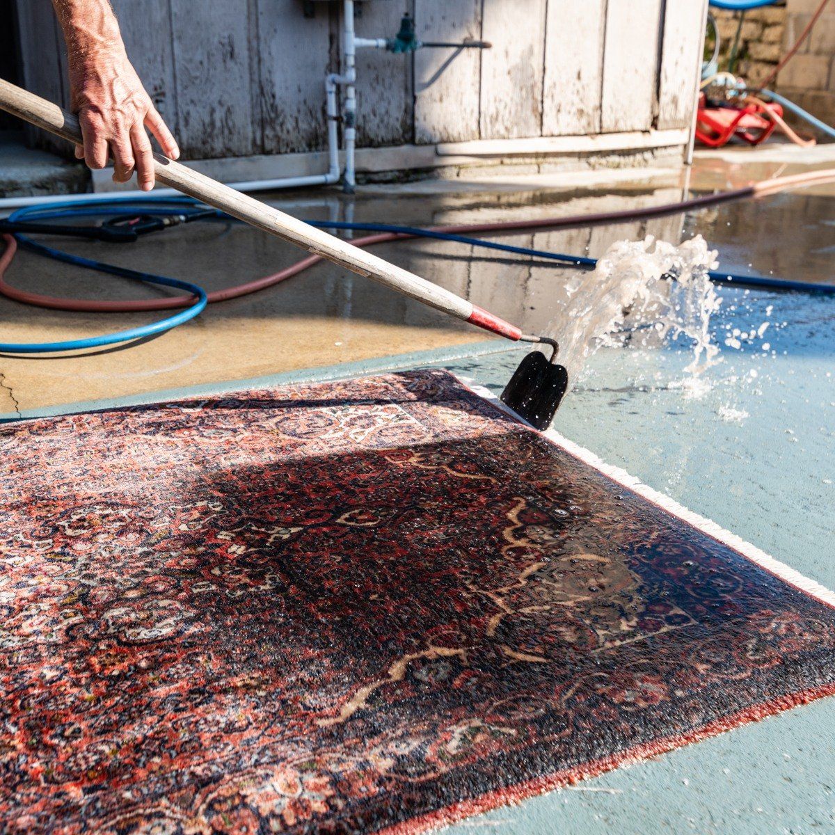 A person uses a pressure washer wand to spray and clean a dark, patterned area rug on a concrete surface.