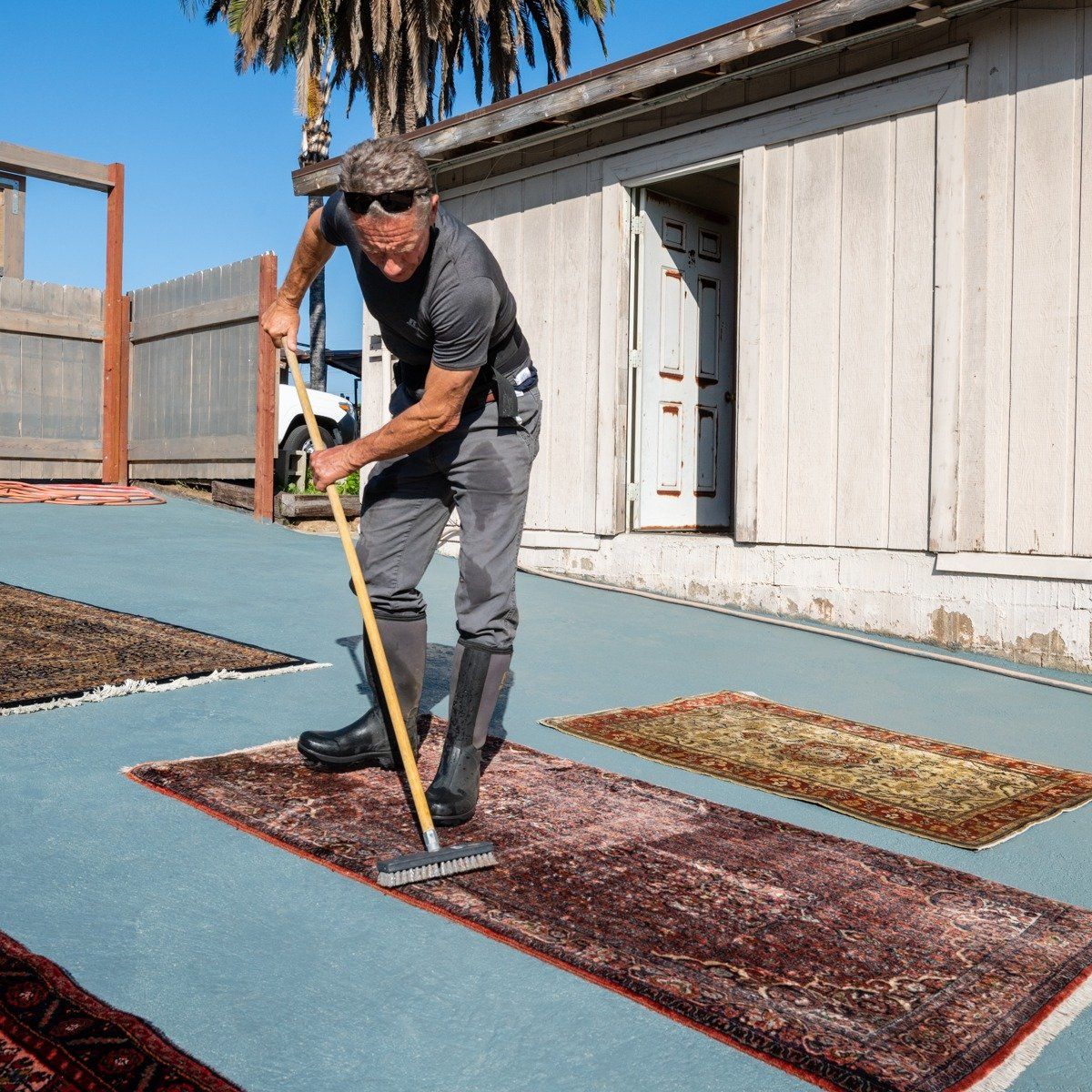 A person in a gray shirt and dark pants scrubs a rug on a paved outdoor surface with a long-handled brush.