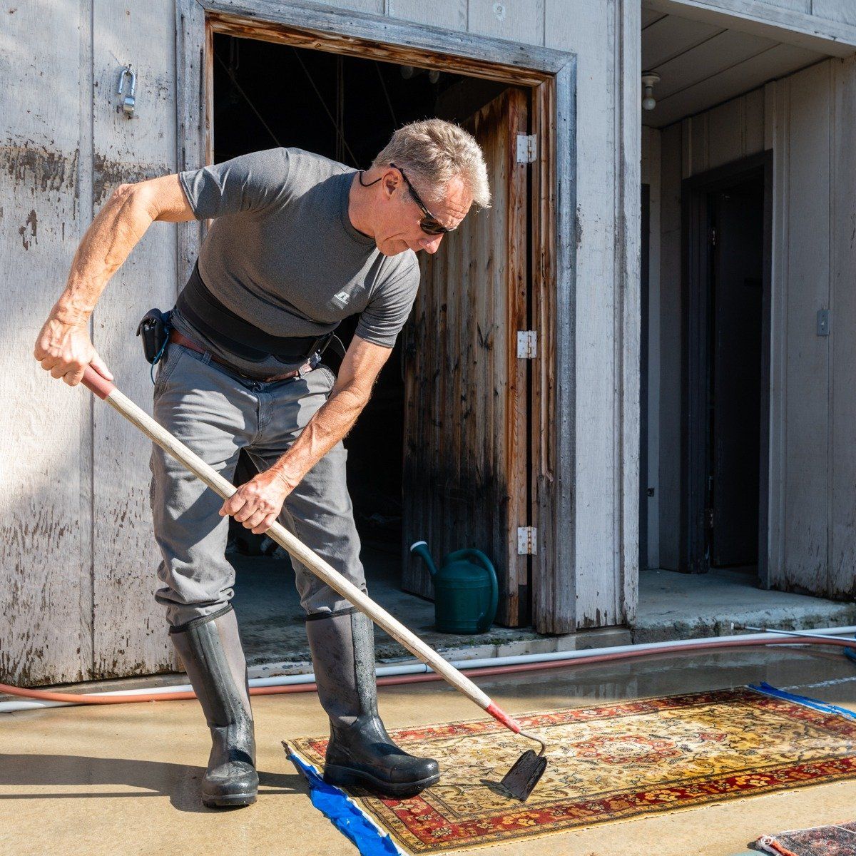 A person in a gray shirt and boots uses a garden hoe to scrub a patterned rug on a concrete patio.
