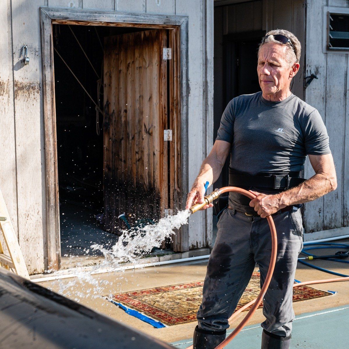 A person in a gray t-shirt and work pants sprays water from a garden hose outside a rustic building.