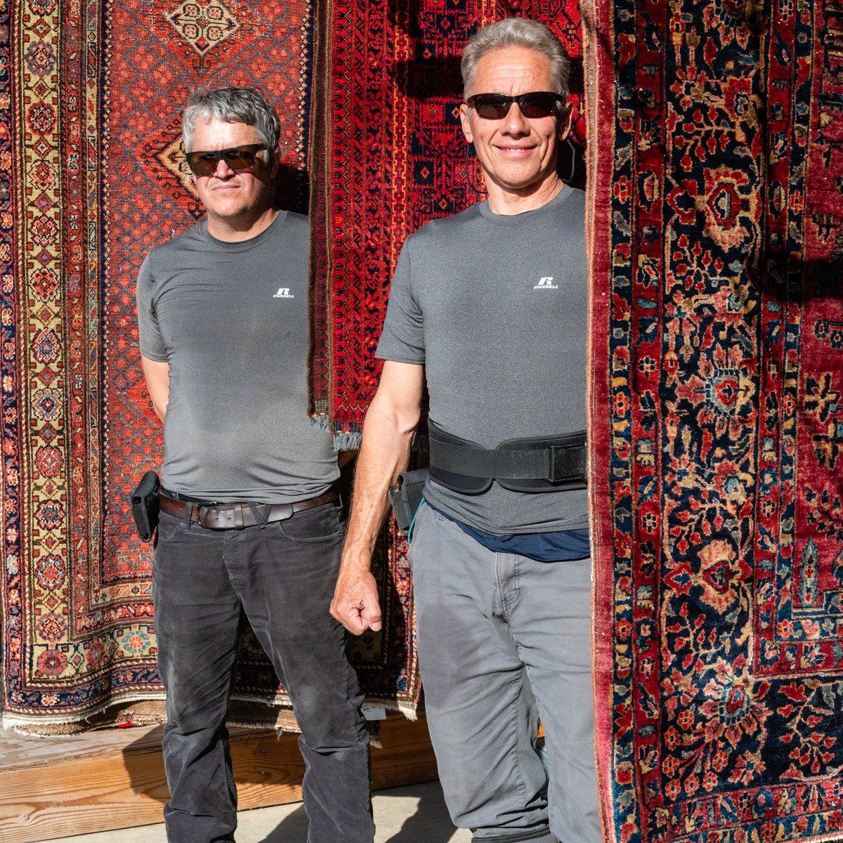 Two men in gray shirts and sunglasses stand in front of several large, intricately patterned, red-toned oriental rugs.
