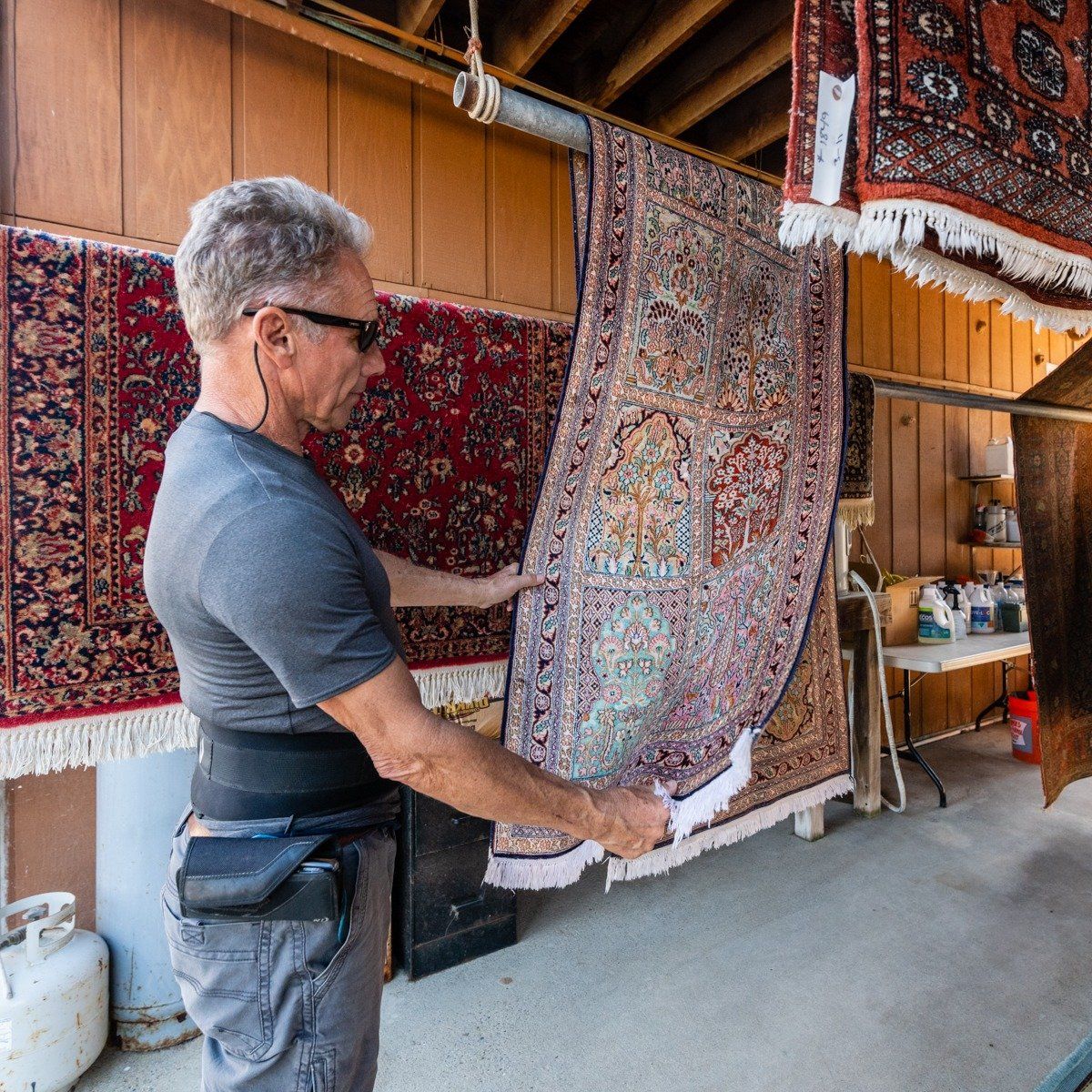 A man wearing a back brace inspects a hanging decorative rug in an indoor workspace filled with other rugs.
