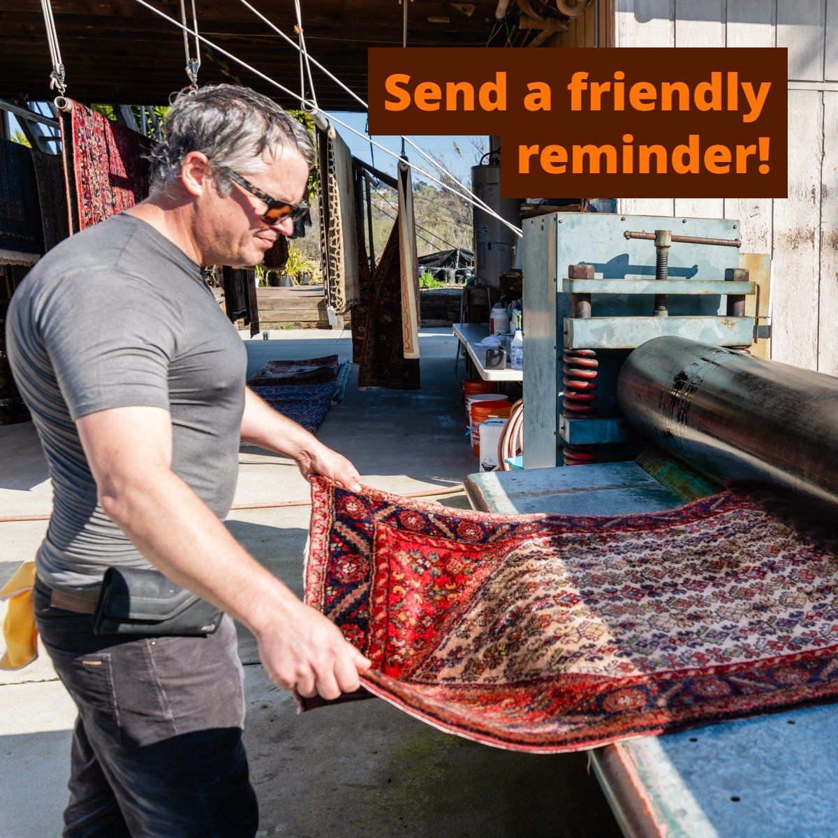 A person wearing sunglasses and a grey t-shirt feeds a colorful patterned rug into a large cleaning or pressing machine.