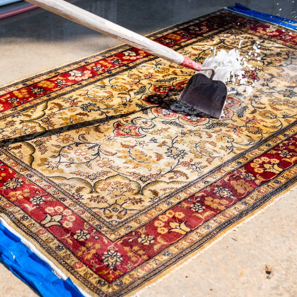 A person uses a squeegee to scrub soapy water across a patterned oriental rug laid out on a concrete surface.