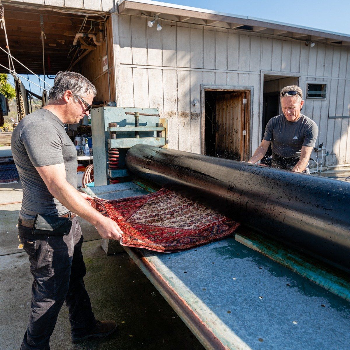 Two people operate a large machine to process a textured, reddish-brown material on a table outside a wooden building.