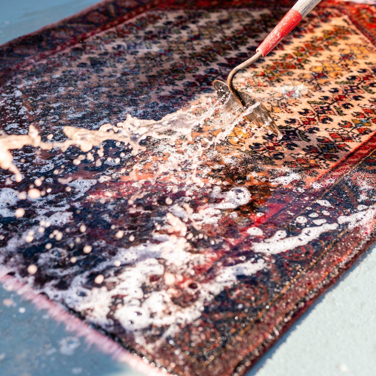 A rug being cleaned by a stream of water from a handheld spray nozzle, creating soapy bubbles on its patterned surface.