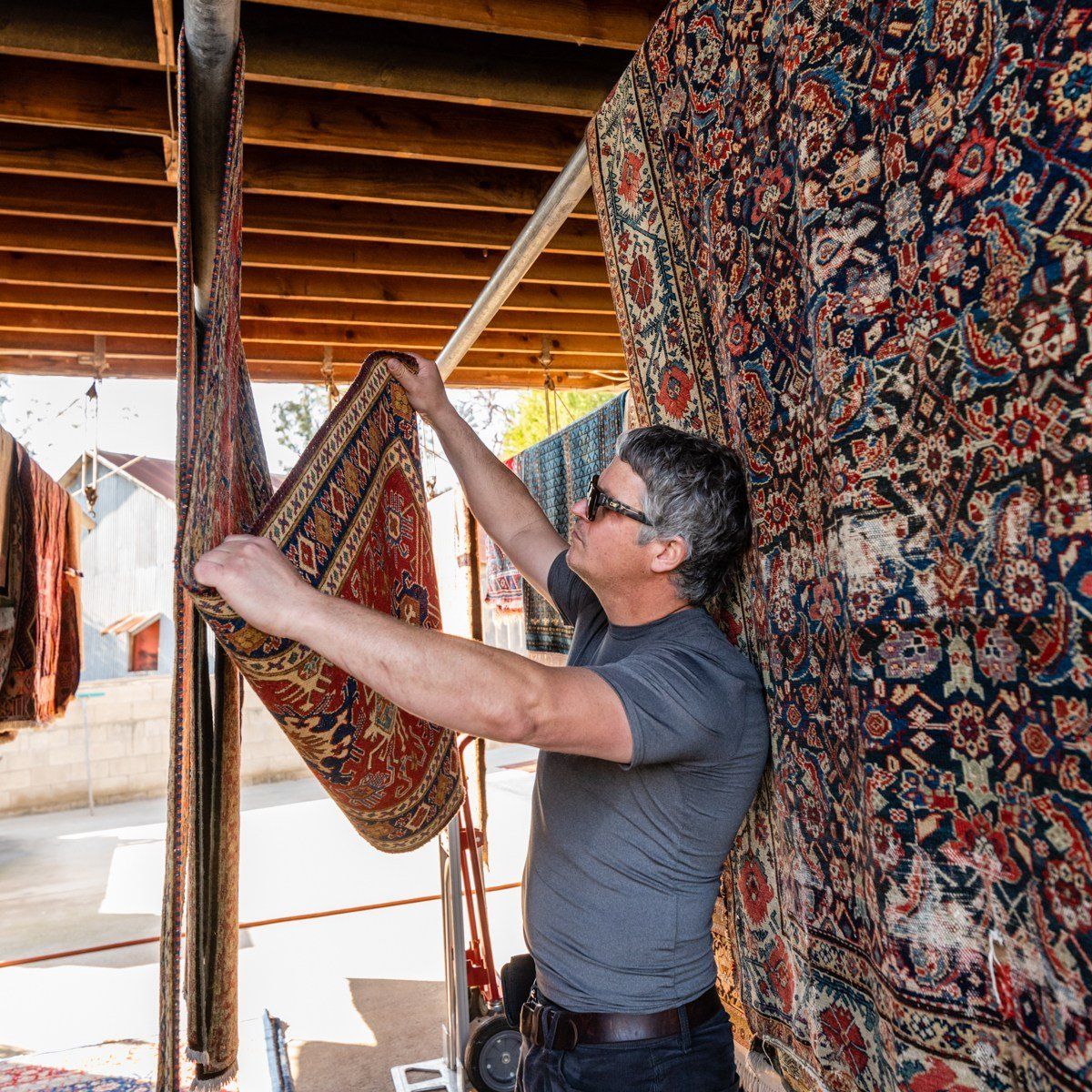 A person hangs an ornate patterned rug on a drying rack under a wooden structure among other hanging carpets.