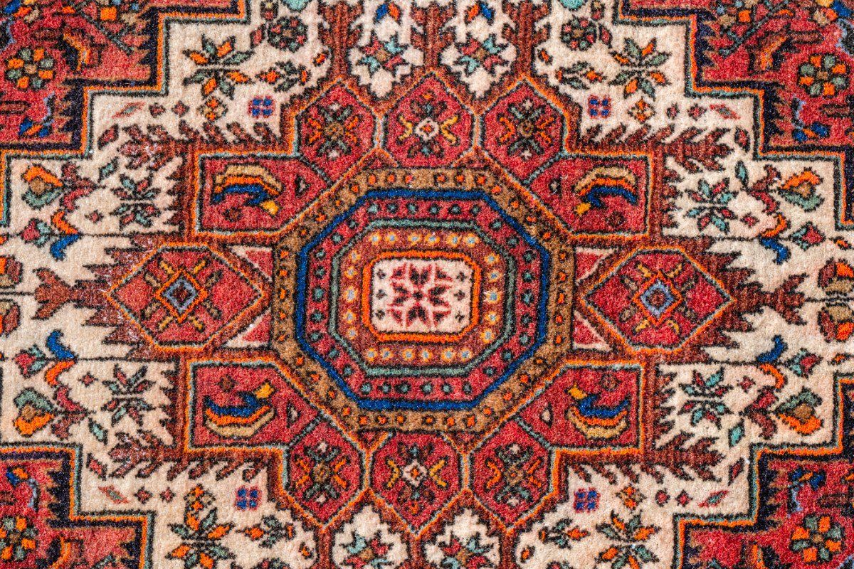 A close-up of a woven rug featuring a symmetrical, geometric medallion pattern in red, blue, cream, and orange hues.