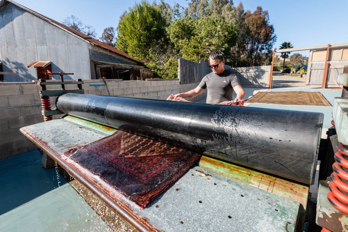 A person operates an industrial printing press with a large black roller over a textured, dark-colored material outdoors.