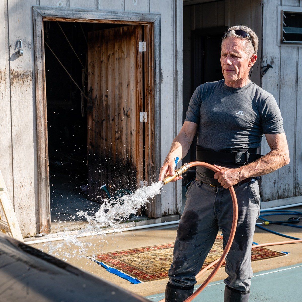 A person in a gray t-shirt and work pants sprays water from a garden hose outside a weathered wooden building.