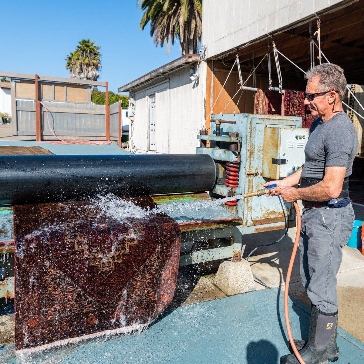 A person uses a hose to spray water on a rug being processed by a large industrial wringer machine outdoors.