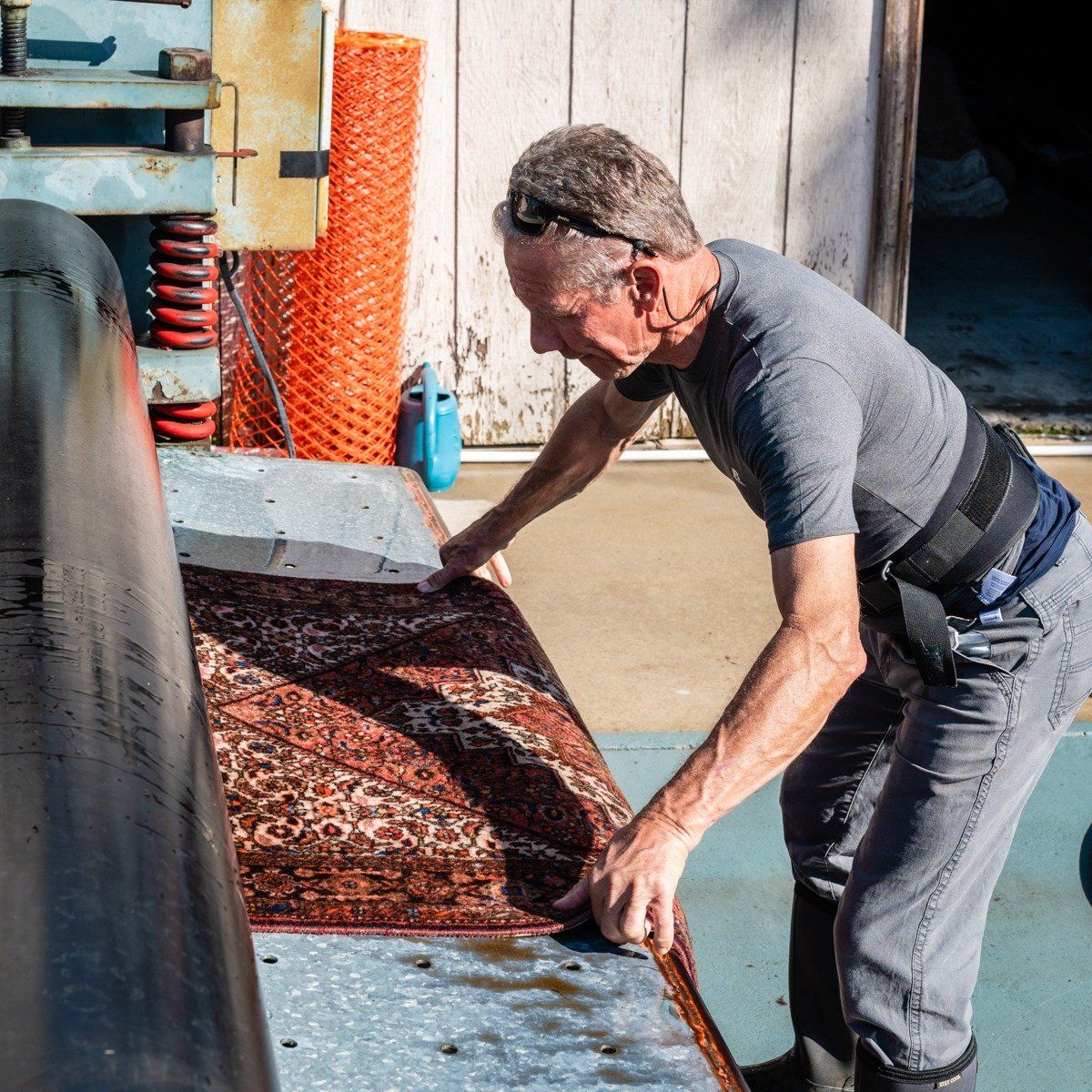 A person in a gray shirt feeds a patterned rug through a large industrial wringer machine at an outdoor wash facility.