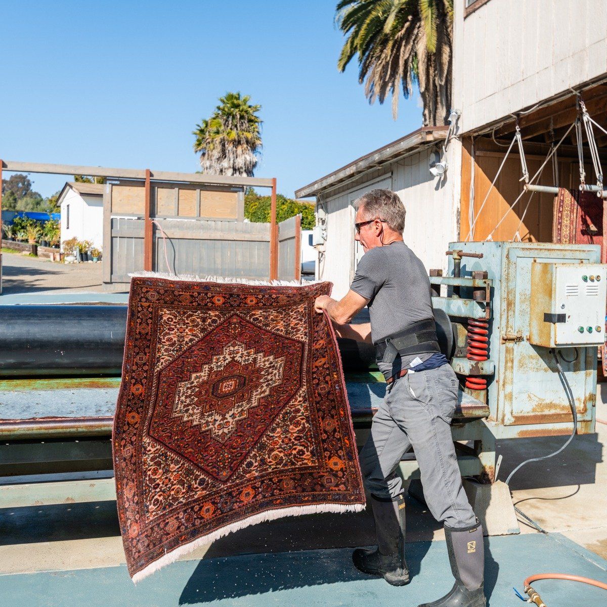 A person in work gear feeds a dark, patterned rug into a large cleaning machine outdoors.