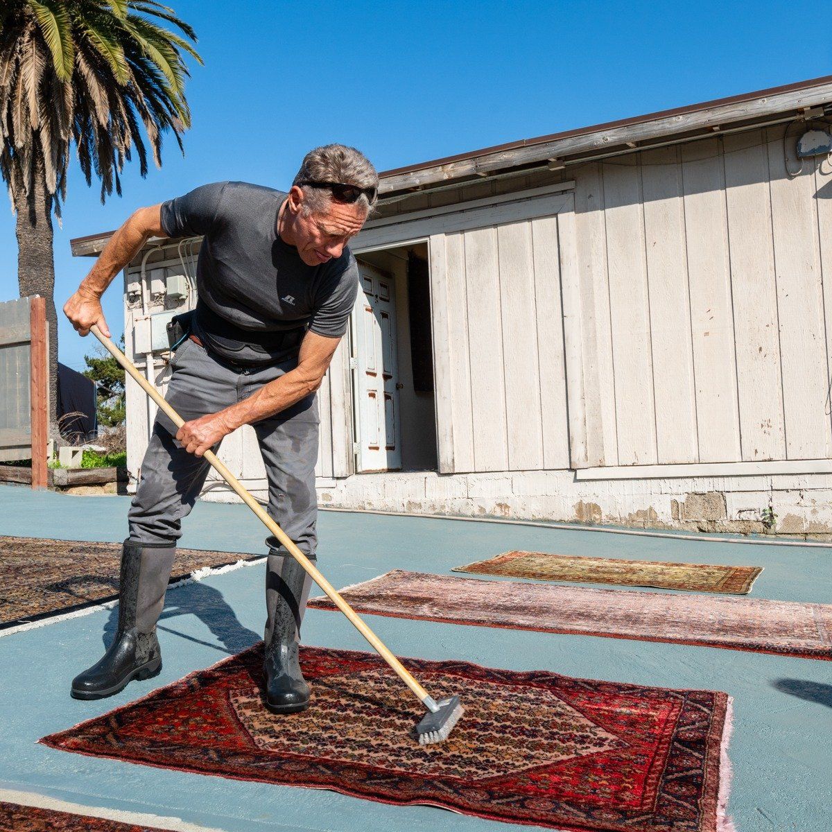 A person wearing a grey shirt and pants uses a long-handled brush to clean an ornate rug on a blue outdoor patio.
