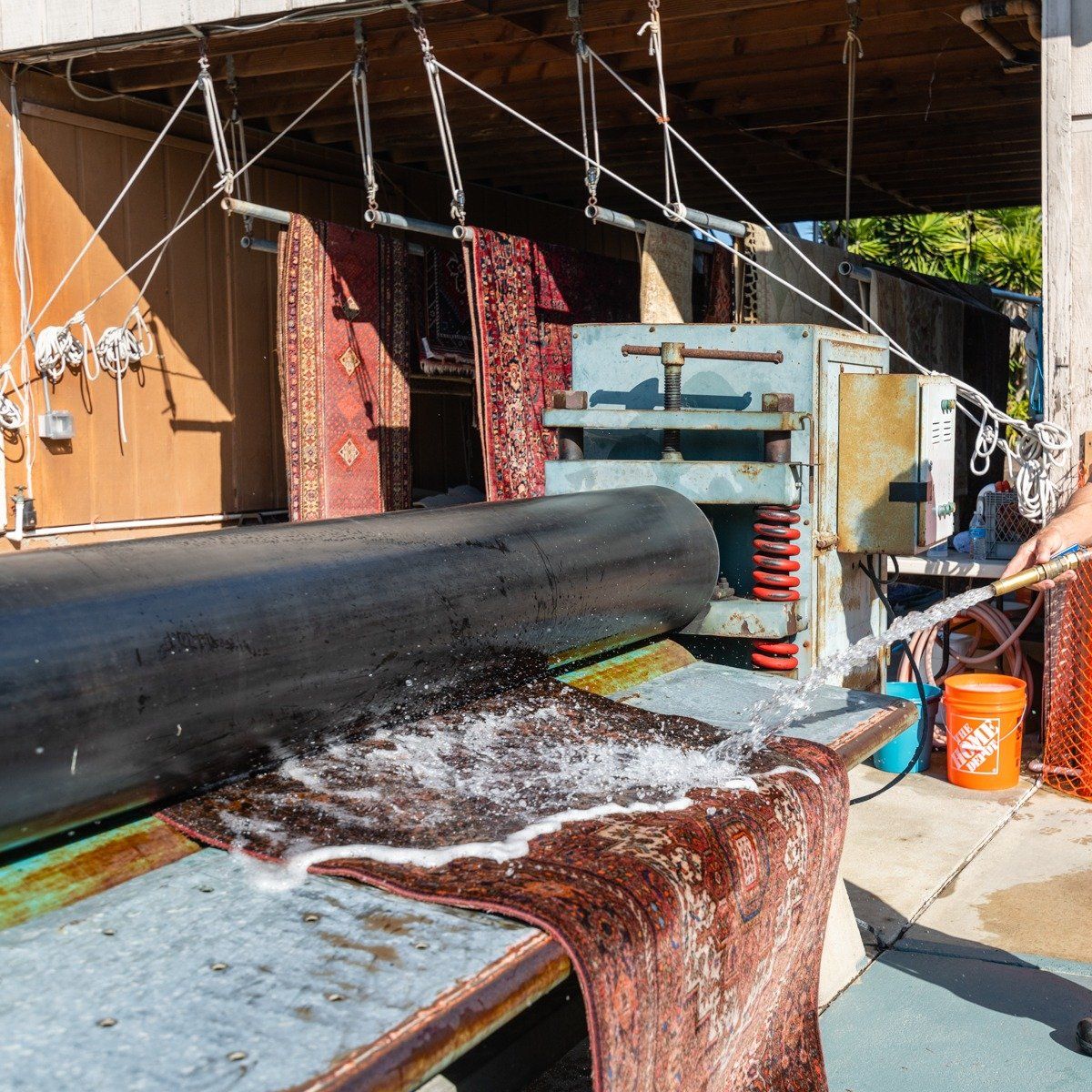 An industrial machine cleans an ornate red rug with pressurized water and rollers in an outdoor workshop.