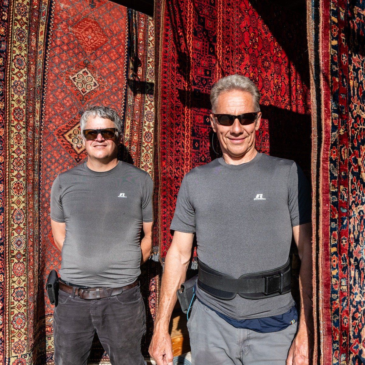 Two people wearing sunglasses and grey t-shirts stand in front of several hanging, intricate patterned rugs.