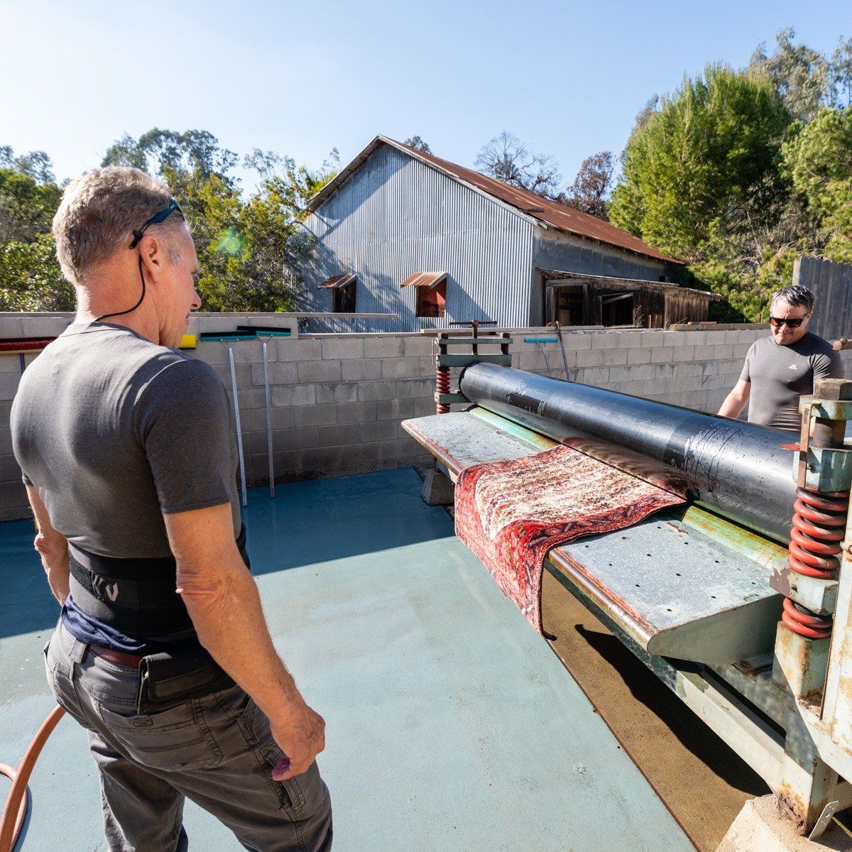 Two people operate a rug-cleaning machine on an outdoor patio with a red patterned rug being rolled through the rollers.