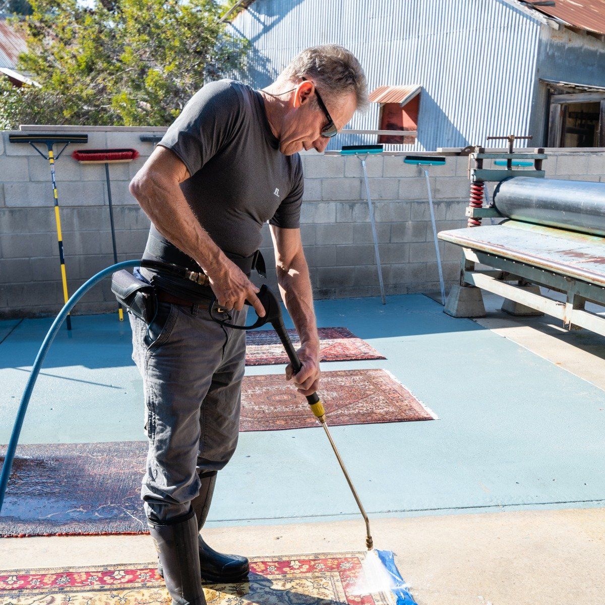 A person uses a pressure washer to clean a rug on a concrete outdoor workspace with several cleaning tools nearby.