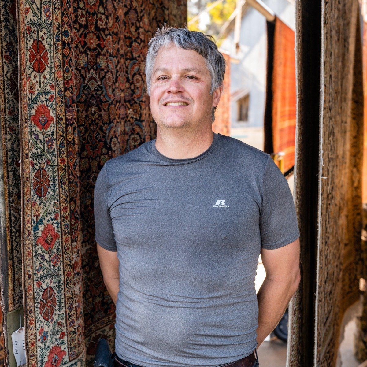 A person with short gray hair smiles, wearing a grey athletic t-shirt while standing between two hanging ornate rugs.