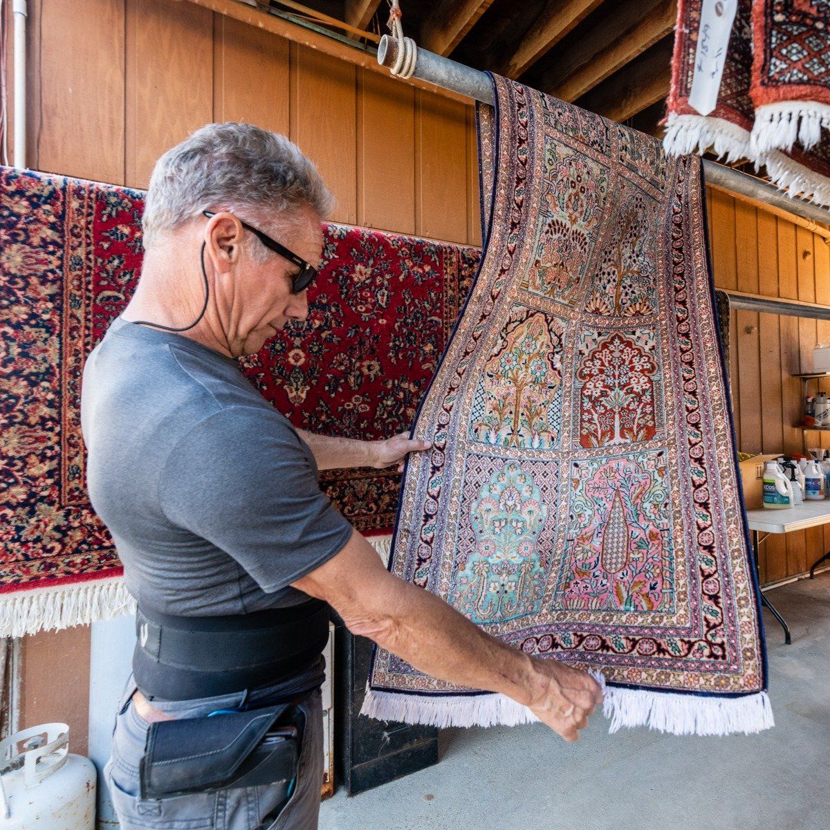 A person wearing a back brace examines a detailed, colorful Persian-style rug hanging in an outdoor storage area.
