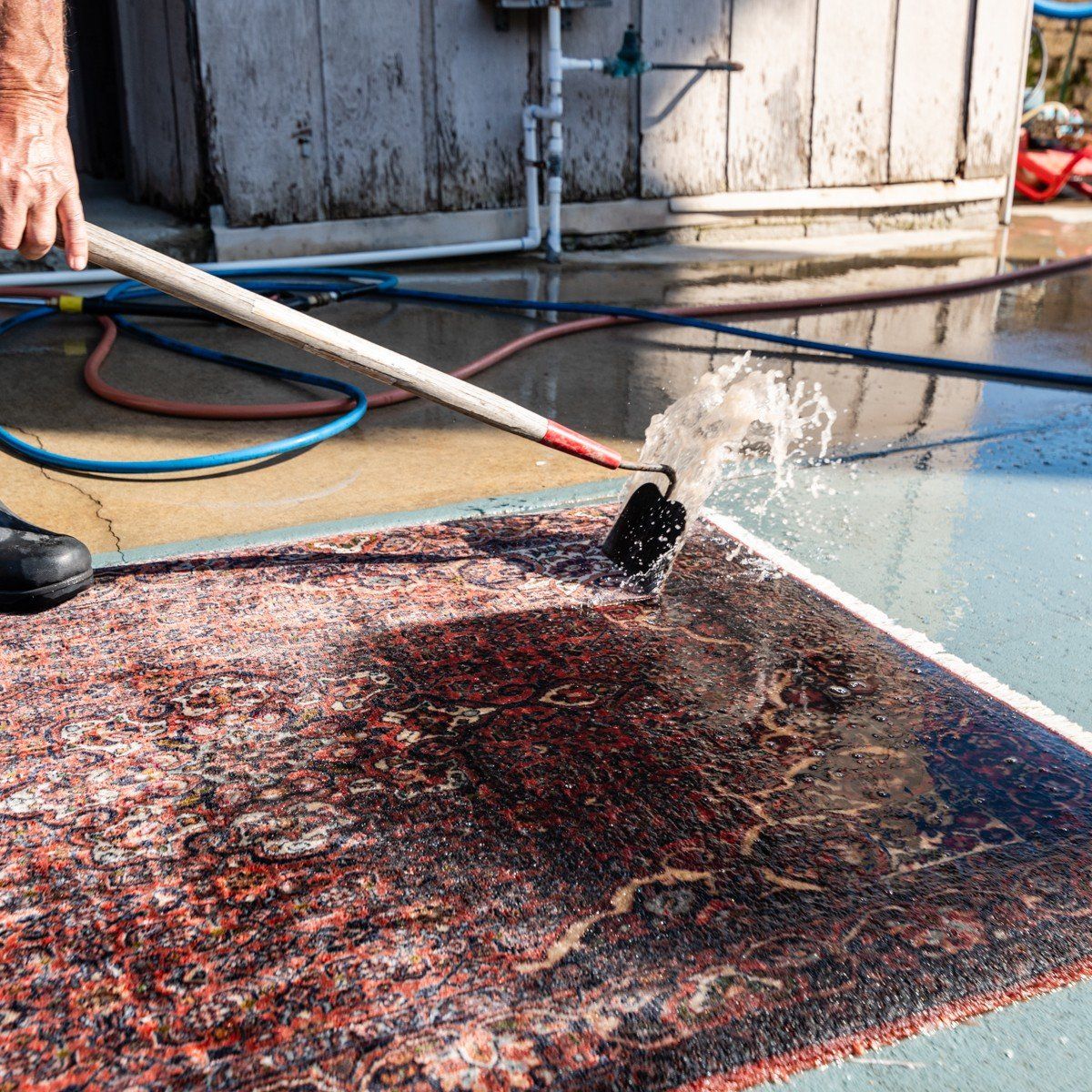 A person uses a handheld squeegee to spray water and clean a patterned rug on a concrete outdoor surface.