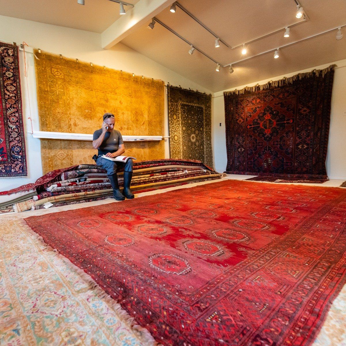 A person sits on stacked carpets in a store showroom filled with numerous large rugs hanging on the walls and floor.