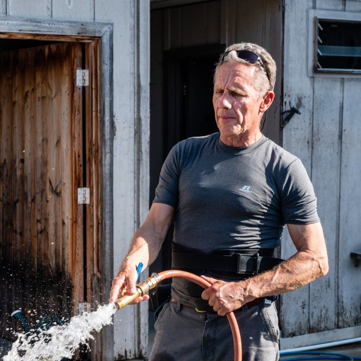 A person in a gray shirt and back brace sprays water from a hose outside a wooden structure.