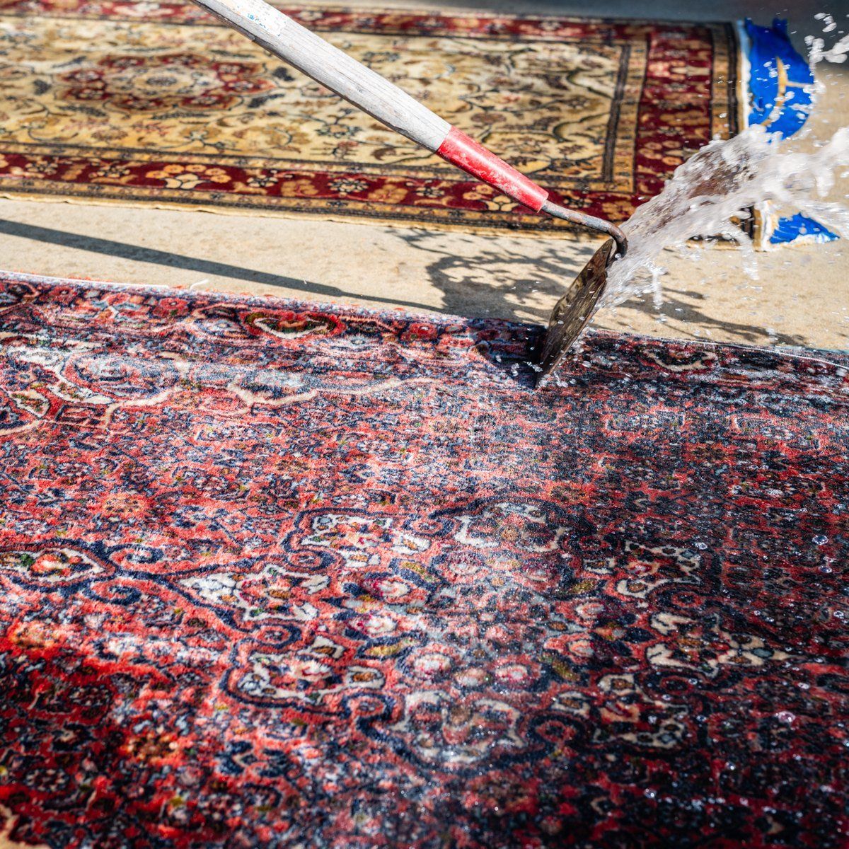 A water hose sprays a patterned, red and blue oriental-style rug outdoors on a concrete surface.