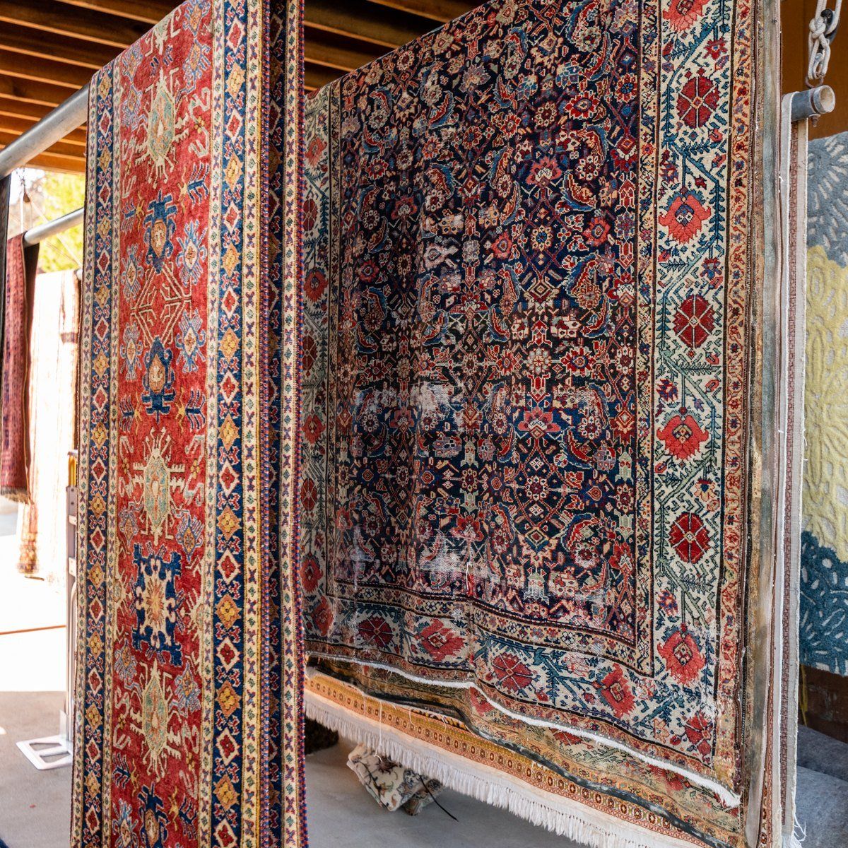Intricately patterned, colorful oriental rugs hang vertically from a metal rack in an indoor display area.