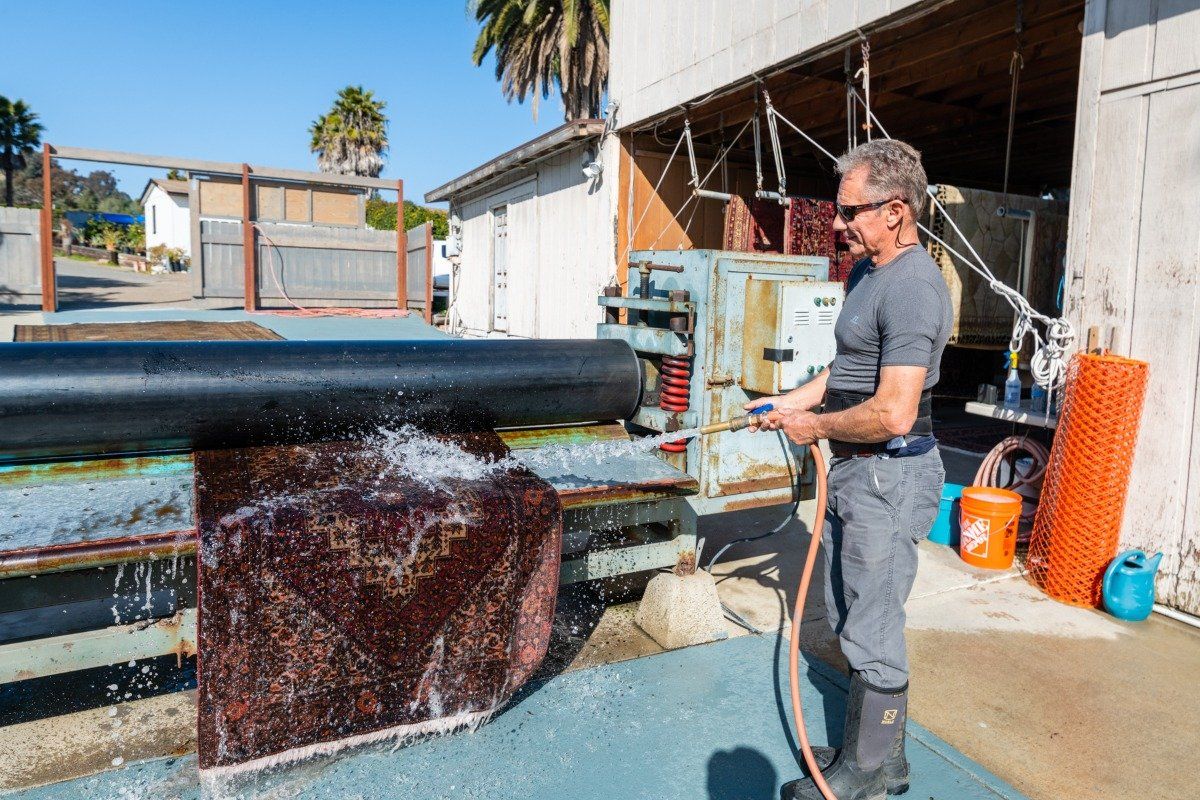 A person in protective boots uses a hose to spray water onto a large dark material in an outdoor industrial area.