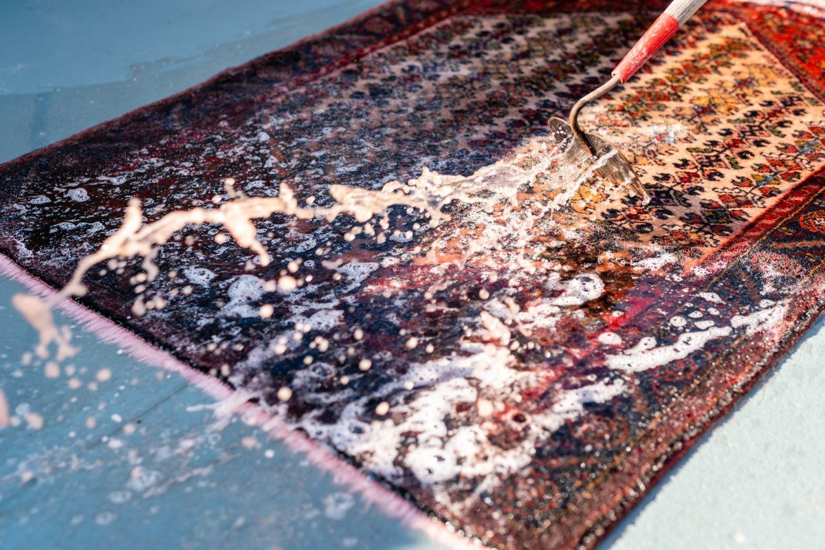 A person uses a handheld tool to spray water and clean a dark, patterned rug lying on a flat surface.