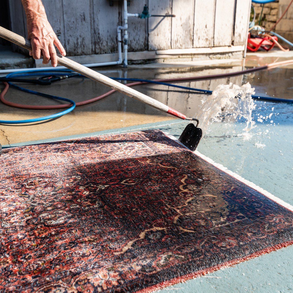 A person uses a pressure washer wand to clean a patterned rug on a concrete surface outdoors.