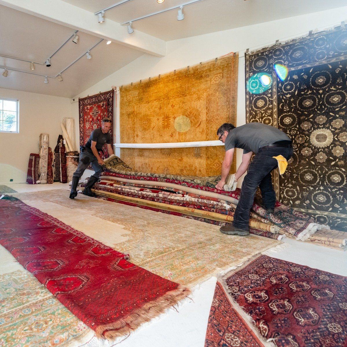 Two people work together to unroll a large, intricately patterned rug in a well-lit showroom filled with area rugs.