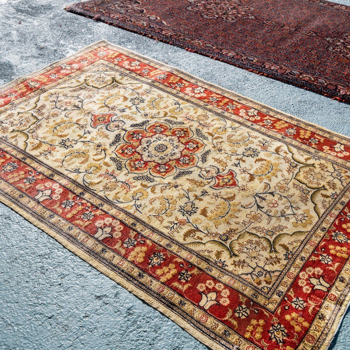 A rectangular rug with a tan center featuring floral motifs and a thick red border, placed on a light grey textured floor.