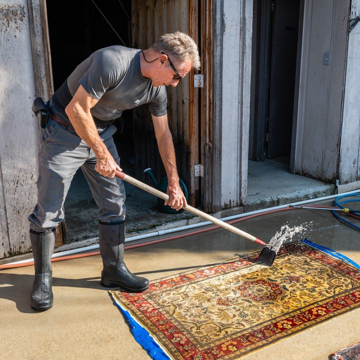A person wearing grey clothing and rubber boots uses a long-handled squeegee to clean a patterned rug on a patio.