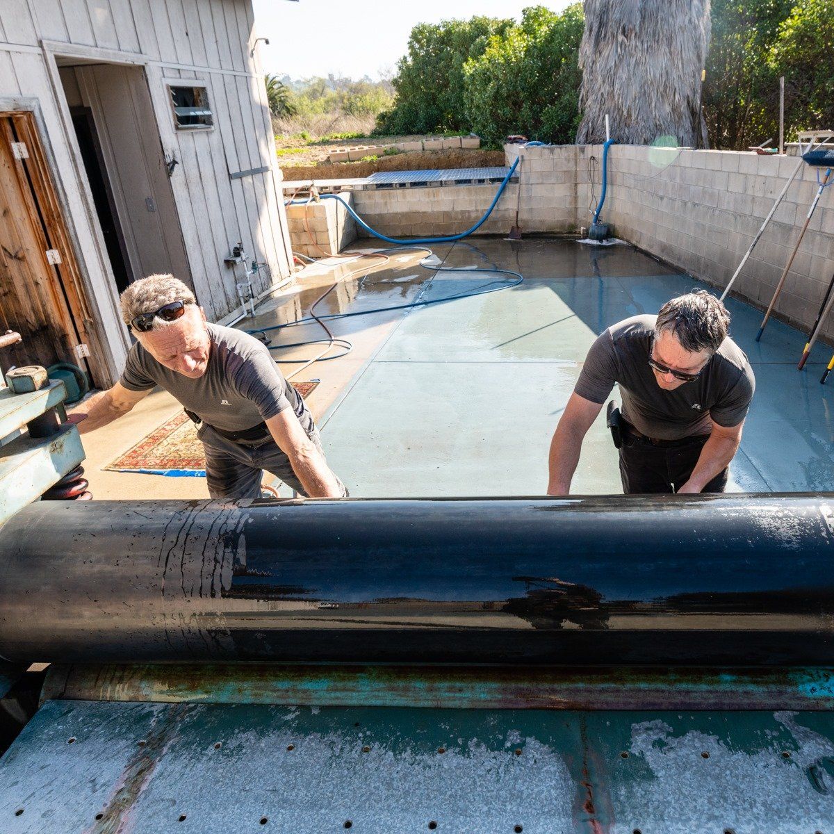 Two workers install a black roofing membrane on a flat roof surface next to a white building.